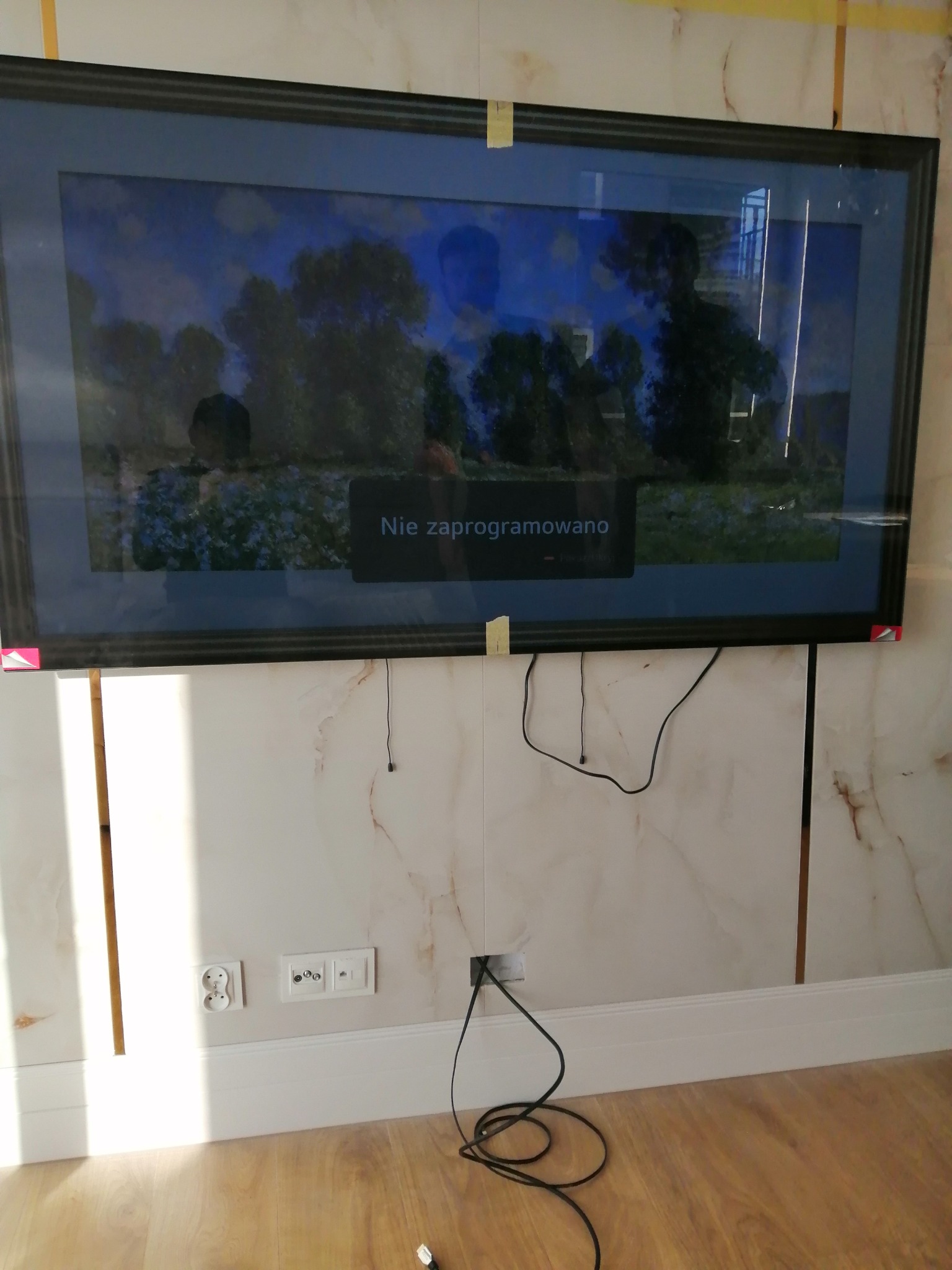 Zamontowany na ścianie wideofon z komunikatem 'Nie zaprogramowano', widoczne kable i gniazdka. Marmurowe wykończenie ściany i drewniana podłoga.