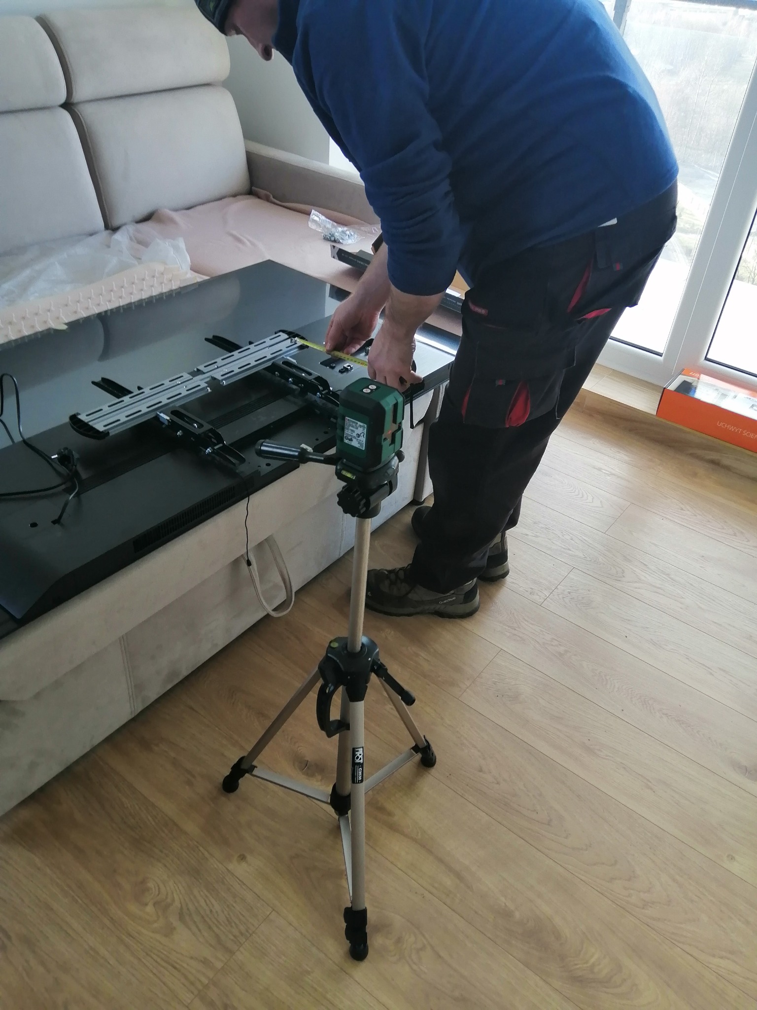 Instalator w trakcie montażu uchwytu ściennego do telewizora, używając poziomicy laserowej na statywie. Widoczny telewizor leżący na kanapie i elementy montażowe.
