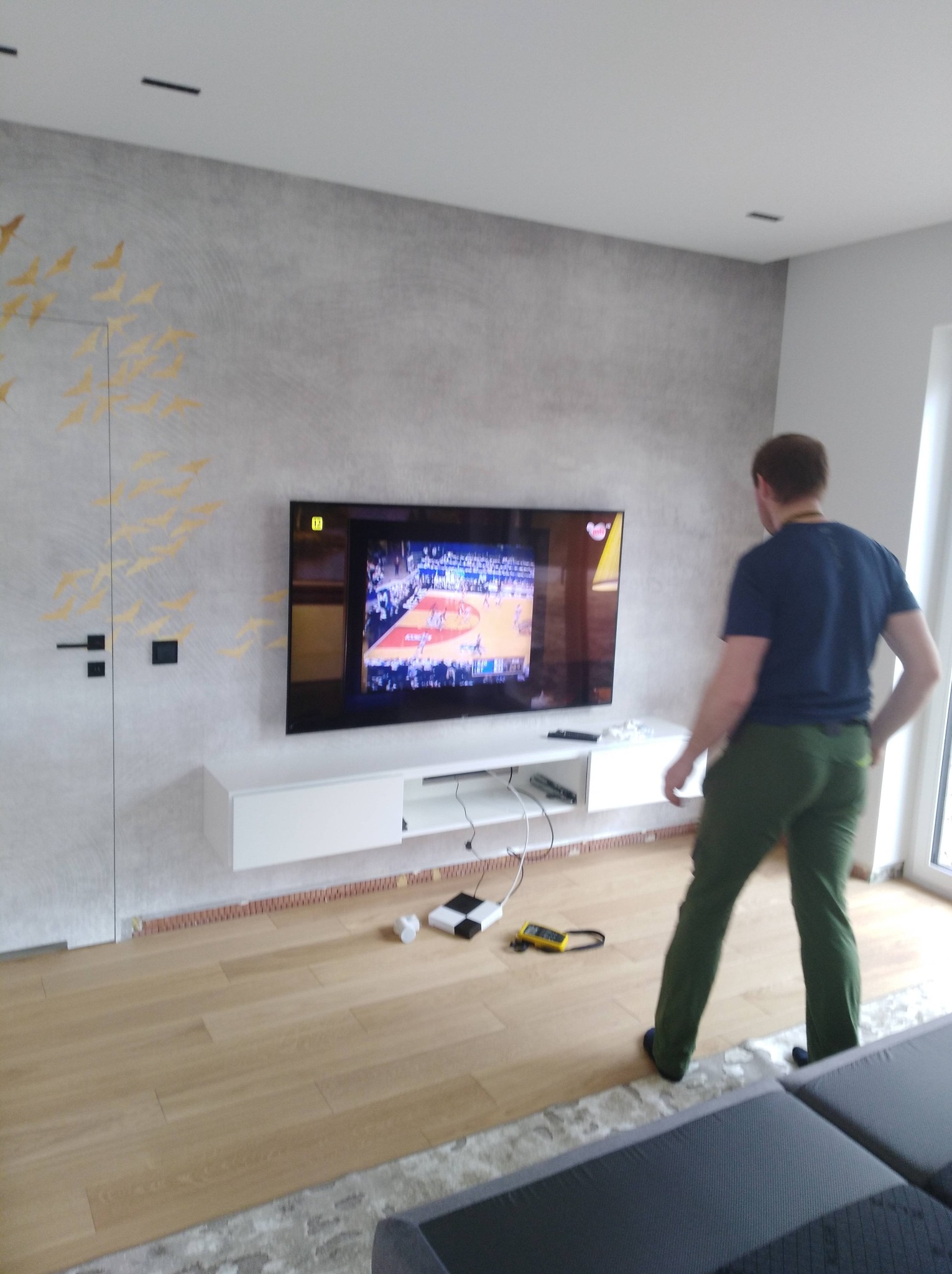 Telewizor na ścianie z podłączonymi kablami i urządzeniami. Widoczny mężczyzna w tle. Instalacja sprzętu RTV w nowoczesnym wnętrzu.