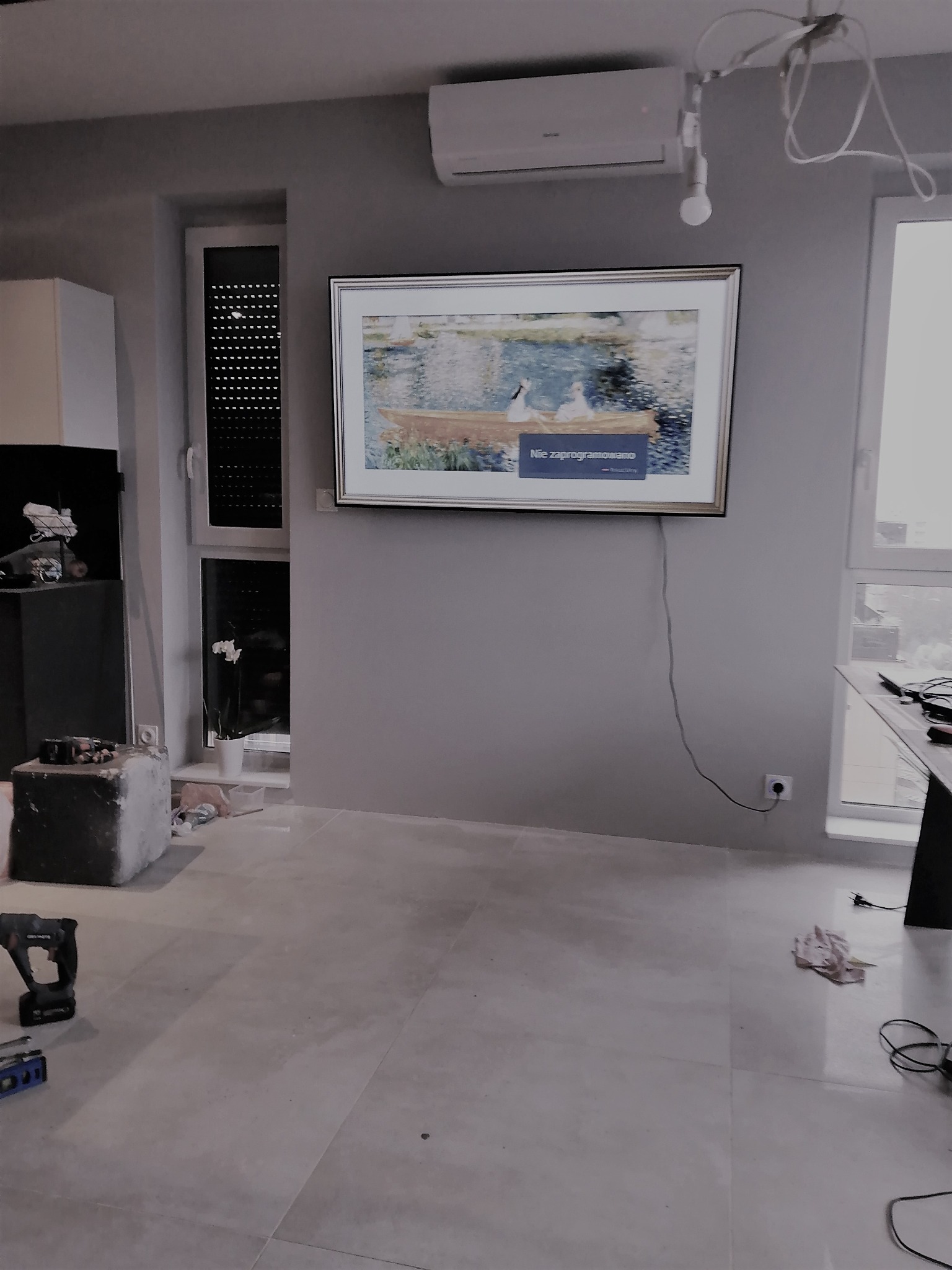 Telewizor z obrazem w ramie zawieszony na szarej ścianie w trakcie remontu. Widoczne narzędzia i materiały budowlane na podłodze.