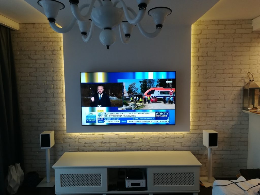 Telewizor na ścianie z cegły, podświetlony LED, z białymi głośnikami po bokach. Biała komoda pod telewizorem. Nowoczesny żyrandol na suficie.