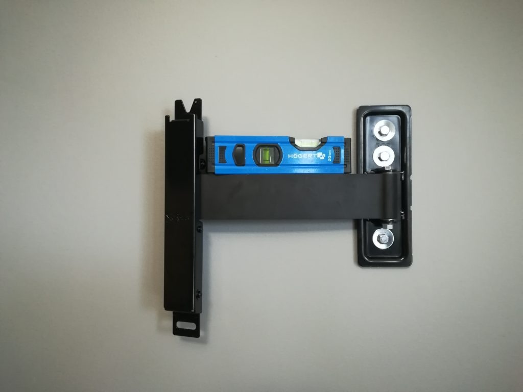 Czarny uchwyt ścienny z poziomicą Högert, zamontowany na szarej ścianie, gotowy do instalacji urządzenia. Widok z przodu, centralne ujęcie.