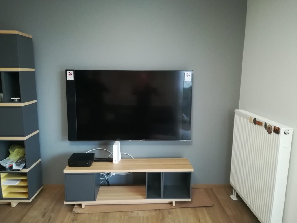 Telewizor na ścianie nad szafką RTV, obok regał i grzejnik. Widoczne kable i urządzenia podłączone do TV. Szaro-beżowa kolorystyka wnętrza.