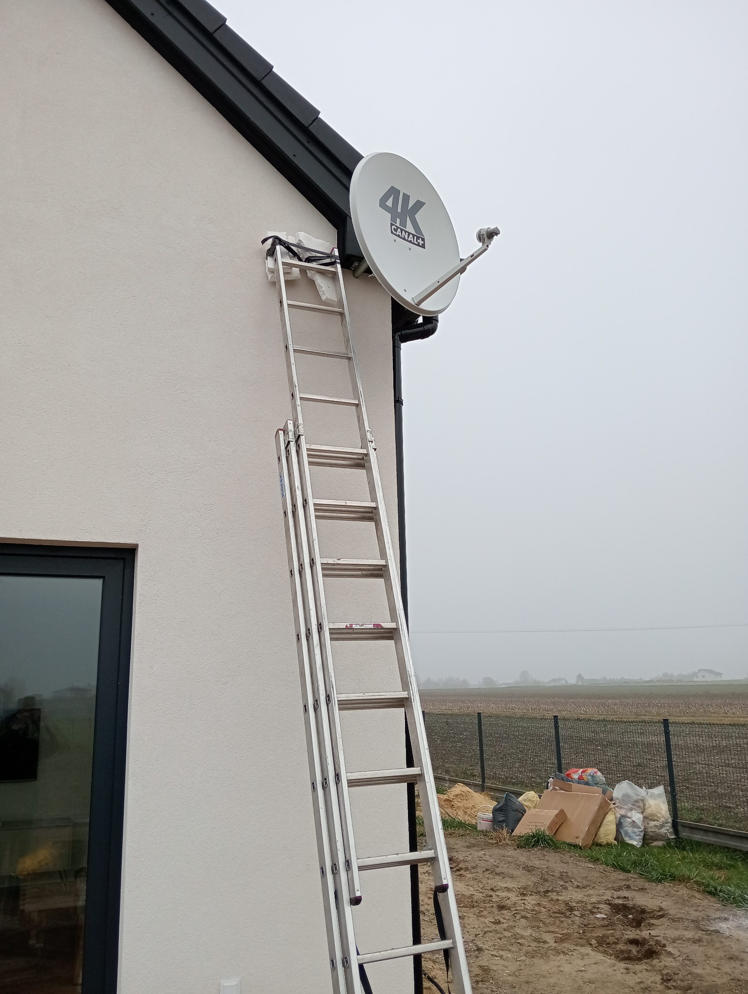 Biała antena satelitarna 4K Canal+ zamontowana na elewacji domu, dostępna po drabinie. Widok na pochmurne pole za ogrodzeniem.