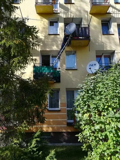 Instalator na drabinie montuje antenę satelitarną na balkonie bloku. Widoczne drzewa i inne anteny. Słoneczny dzień.