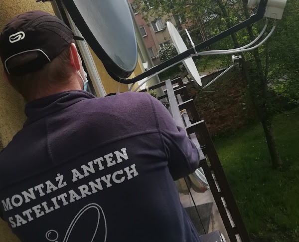 Pracownik w bluzie z napisem 'Montaż Anten Satelitarnych' instaluje antenę satelitarną na balkonie. Widoczne kable i uchwyty montażowe.