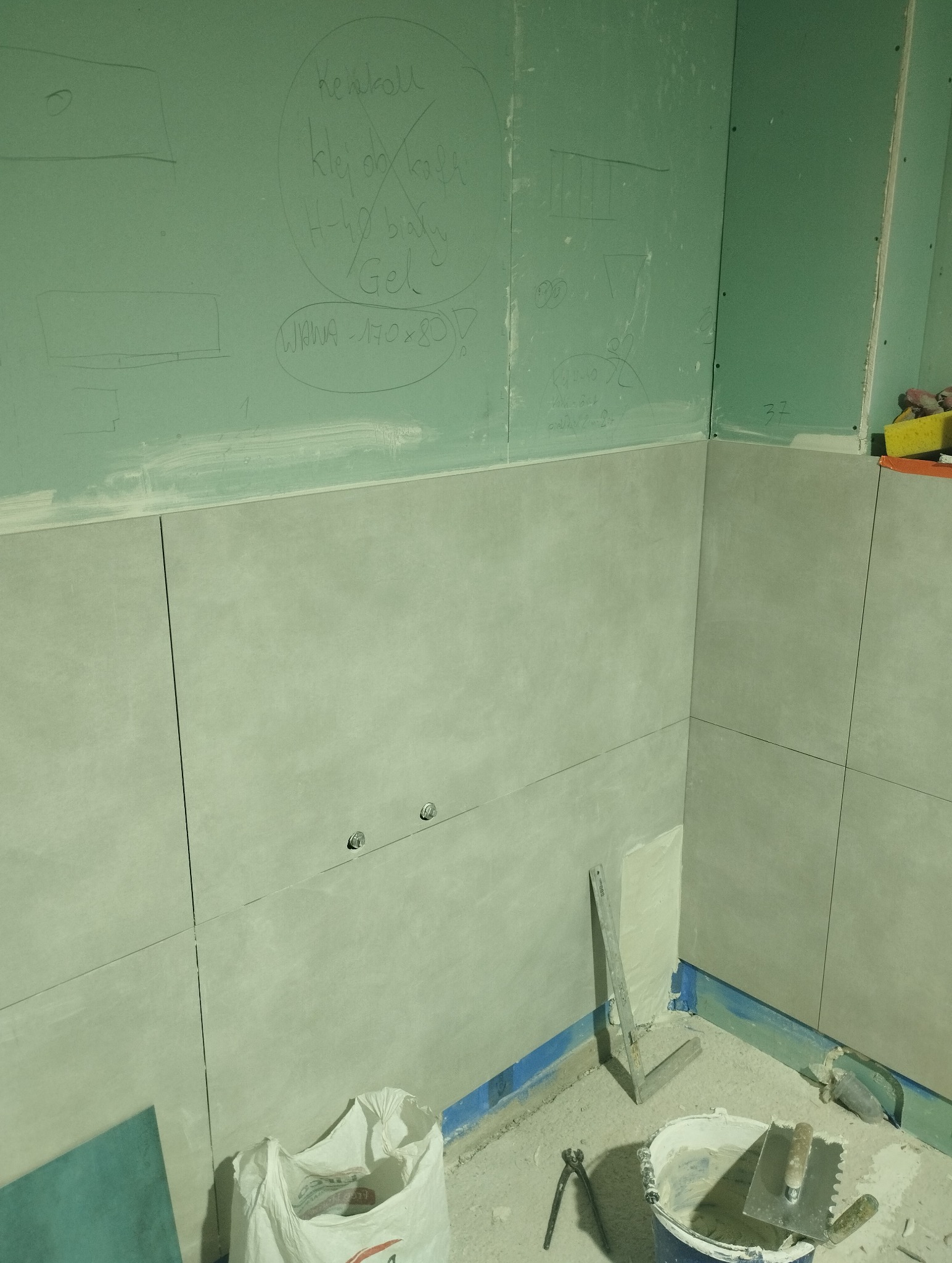 Ściana łazienki w trakcie wykańczania: szare płytki częściowo ułożone, widoczne narzędzia, worek z klejem i notatki na płycie karton-gips.