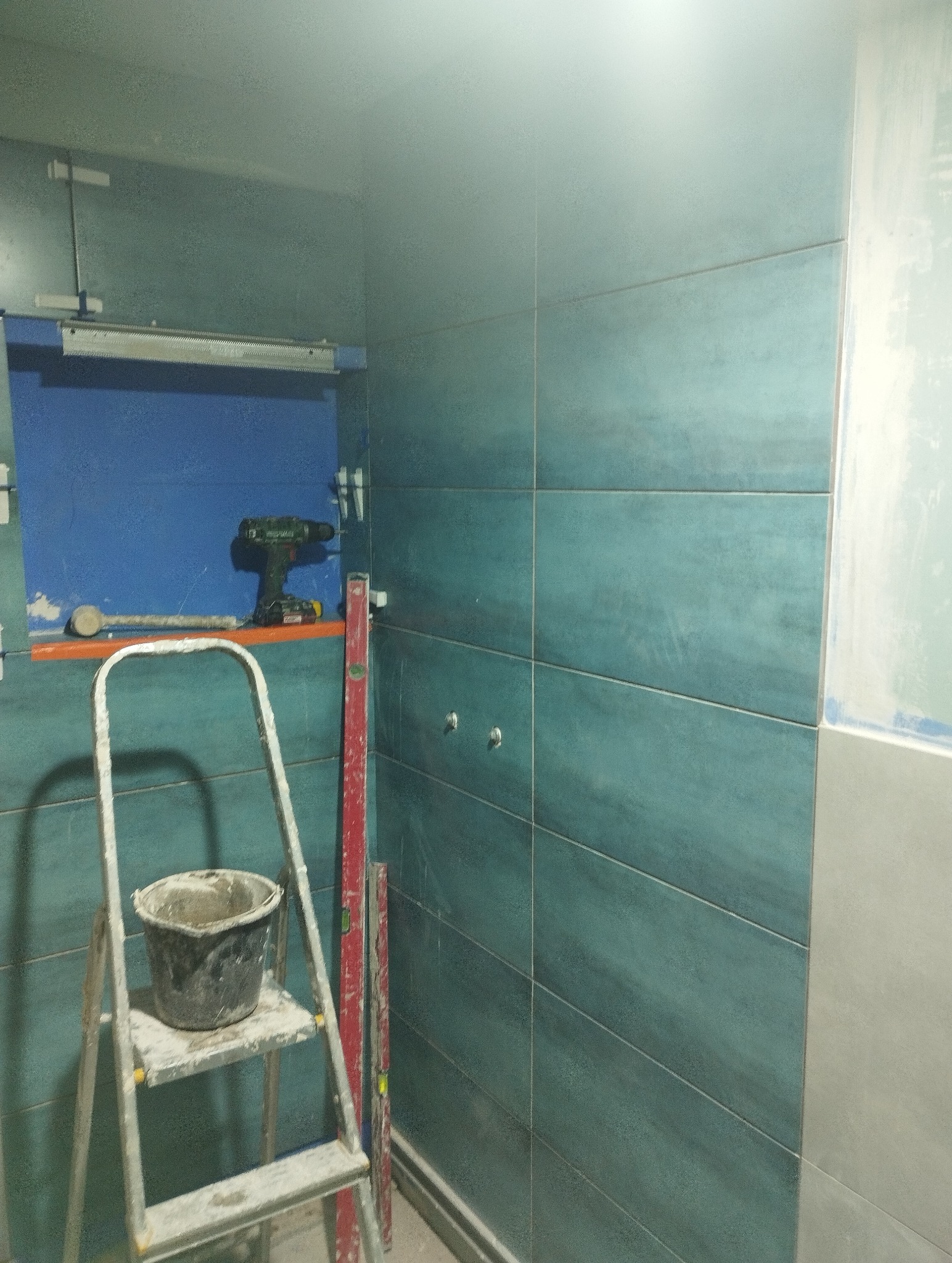 Turkusowe płytki w łazience, drabina z wiadrem, poziomica i wkrętarka w niebieskiej wnęce. Etap prac wykończeniowych, widoczne fugi i fragment surowej ściany.