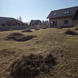 USŁUGI OGRODNICZE MARCIN BABIŃSKI - Teren po wycince krzewów z widocznymi kupkami gałęzi i częściowo wykarczowaną ziemią. W tle dom w trakcie budowy i zabudowa jednorodzinna.