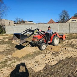 USŁUGI OGRODNICZE MARCIN BABIŃSKI - Czerwony traktor z łyżką na polu z piaskiem i resztkami trawy, operator w szarej bluzie. W tle betonowe ogrodzenie i budynki pod błękitnym niebem.