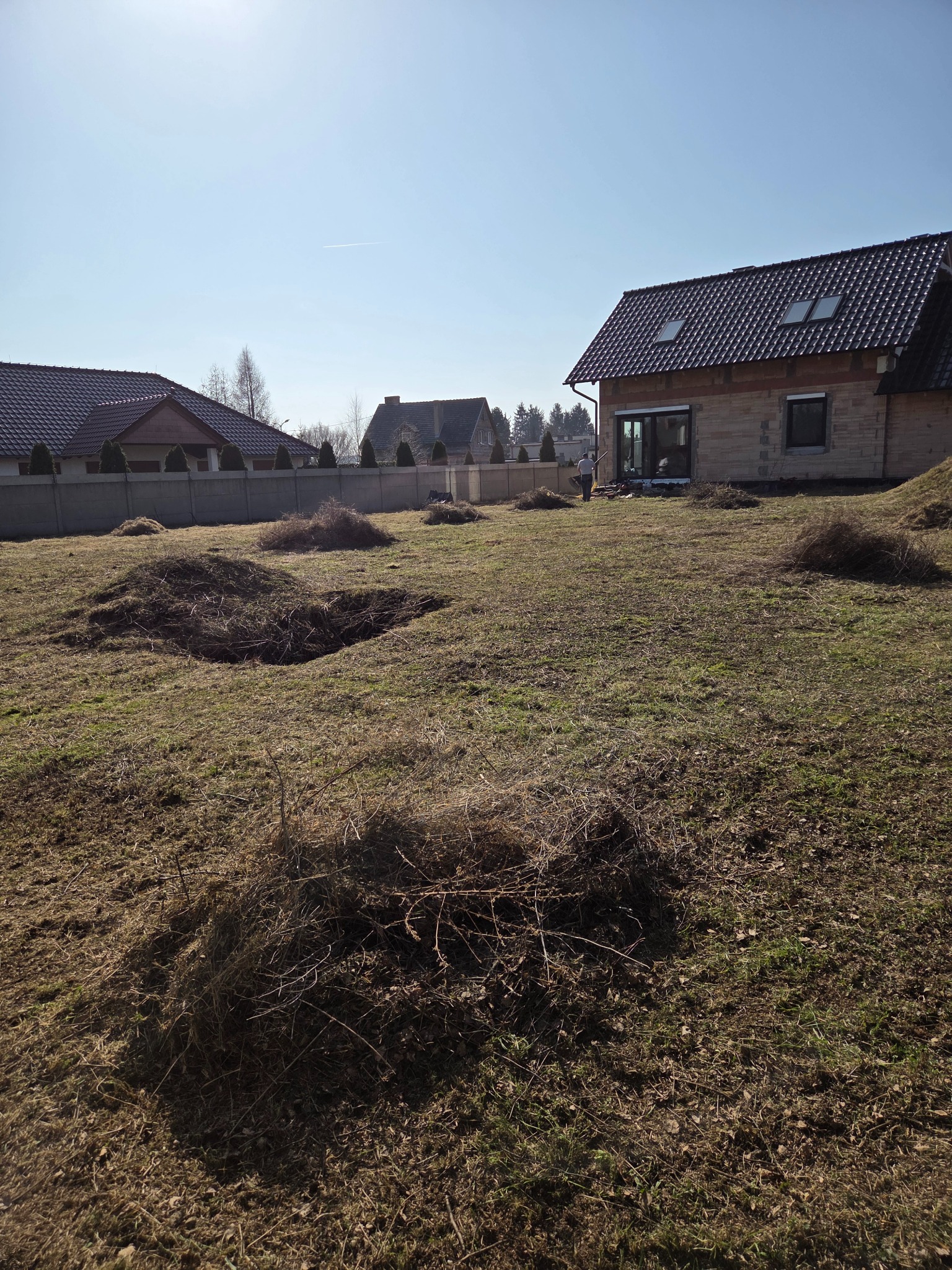 Teren po wycince krzewów z widocznymi kupkami gałęzi i częściowo wykarczowaną ziemią. W tle dom w trakcie budowy i zabudowa jednorodzinna.