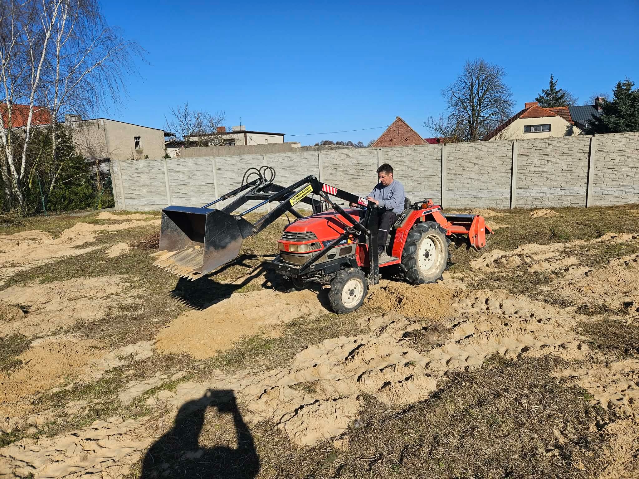 Czerwony traktor z łyżką na polu z piaskiem i resztkami trawy, operator w szarej bluzie. W tle betonowe ogrodzenie i budynki pod błękitnym niebem.