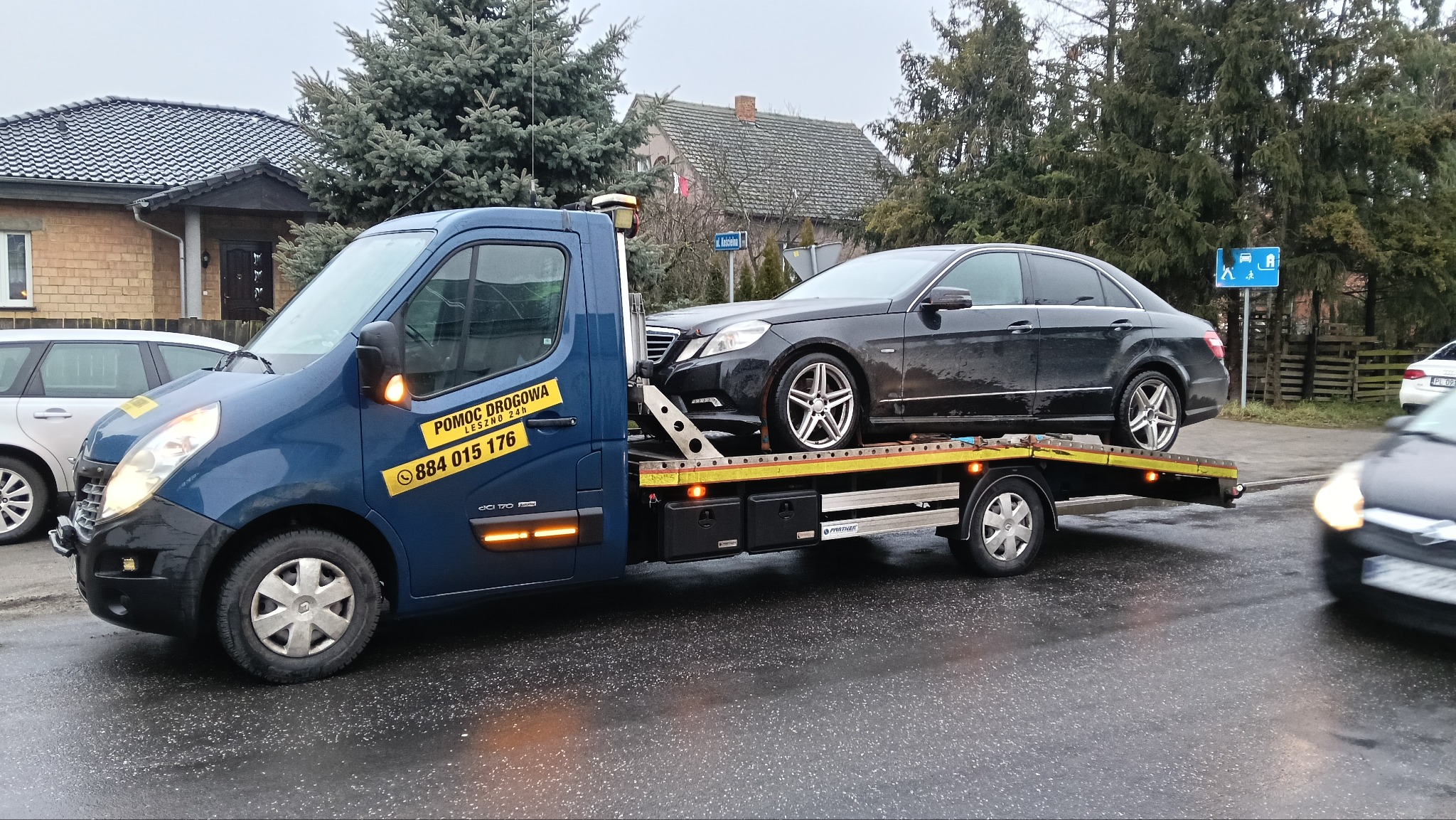 Niebieska laweta z czarnym Mercedesem na platformie, widoczna naklejka z numerem telefonu. Tło: zabudowa miejska, pochmurny dzień.