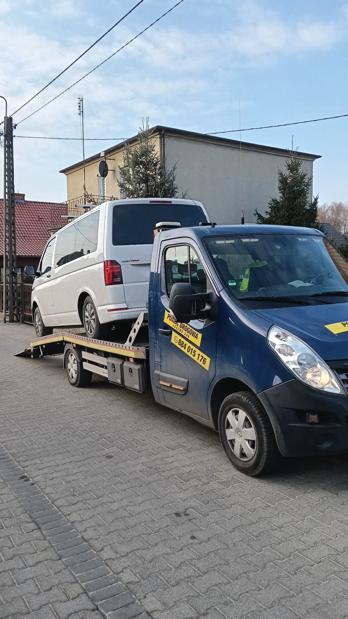 Niebieska laweta z białym vanem na platformie, zaparkowana na brukowanej ulicy w Lesznie, z naklejką kontaktową 'Pomoc Drogowa Leszno 24h' na drzwiach.