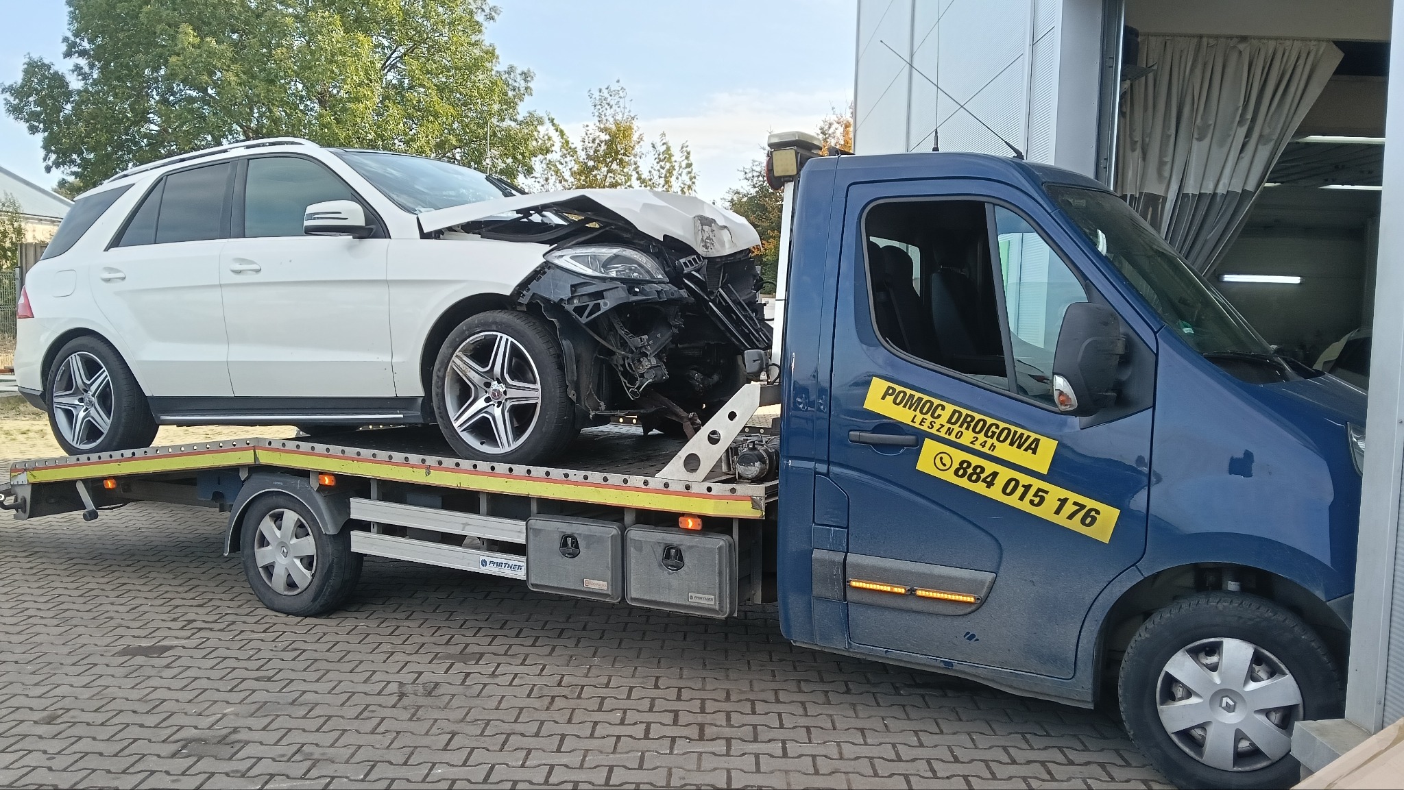 Niebieska laweta z uszkodzonym białym SUV-em Mercedes na platformie. Widoczny numer telefonu pomocy drogowej Leszno. Platforma lawety z żółtymi światłami.