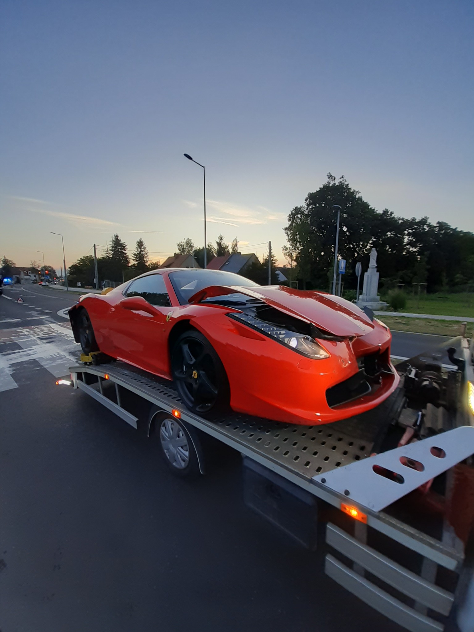 Uszkodzone, czerwone Ferrari na platformie lawety, widoczne uszkodzenia przodu pojazdu. W tle droga, drzewa i budynki. Pomoc drogowa w akcji.