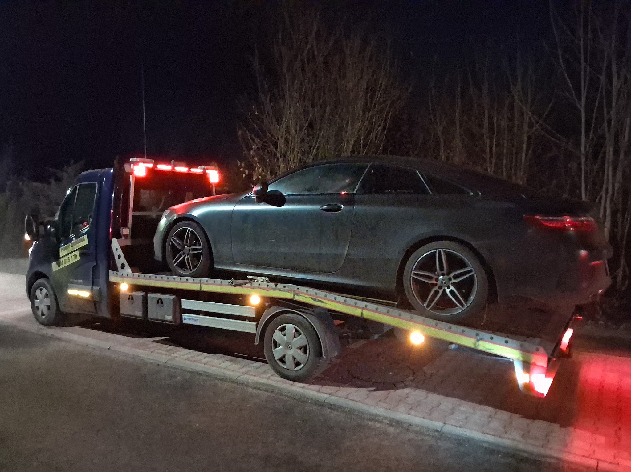 Nocna laweta z Mercedesem E-klasą Coupe na platformie. Widoczne światła ostrzegawcze i numer telefonu pomocy drogowej. Tło stanowią drzewa.