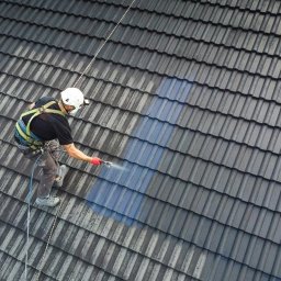 Pracownik w kasku i uprzęży, z góry, czyści ciemny dach z dachówki za pomocą myjki ciśnieniowej. Widoczna różnica czystości dachu.