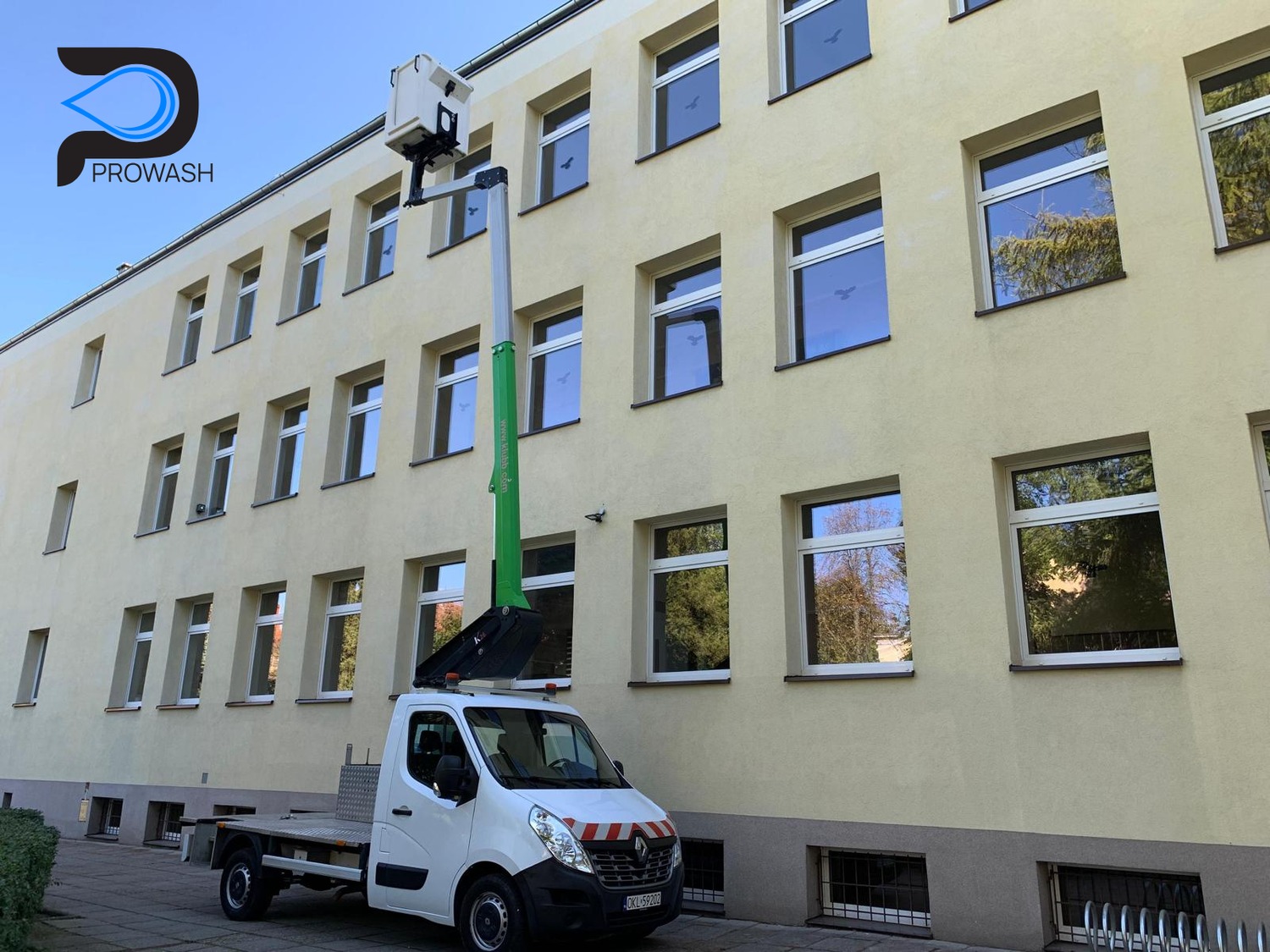 Żółta elewacja budynku z wieloma oknami, czyszczona z podnośnika koszowego na białym samochodzie dostawczym. Widoczne odbicia drzew w szybach okien.