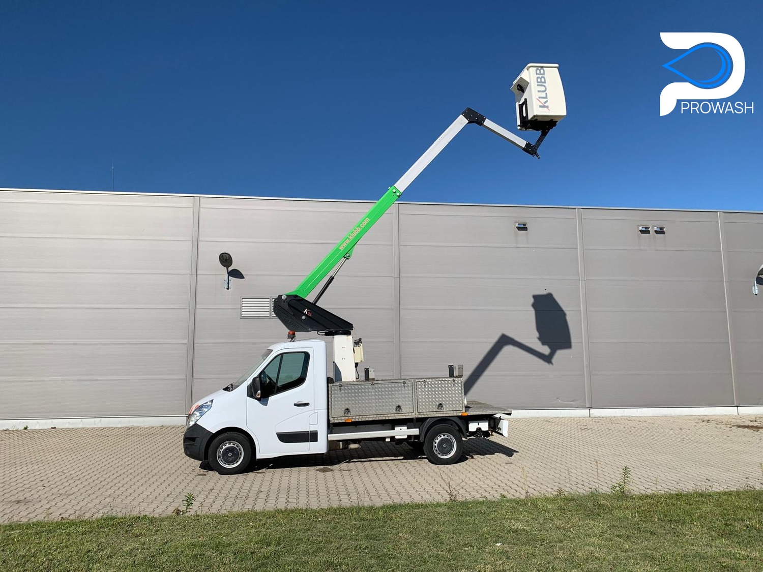 Biały samochód z podnośnikiem koszowym marki Klubb na tle szarej hali przemysłowej i błękitnego nieba. Logo firmy Prowash w prawym górnym rogu.