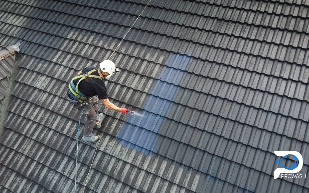 Pracownik w kasku i uprzęży, z góry, czyści ciemny dach z dachówki za pomocą myjki ciśnieniowej. Widoczna różnica czystości dachu.