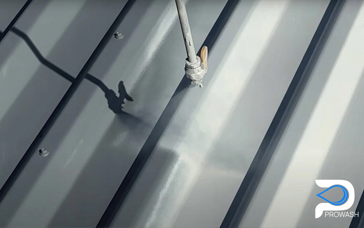 Szare blaszane pokrycie dachowe z dyszą ciśnieniową w trakcie czyszczenia. Widoczny cień węża i dyszy. Logo firmy w prawym dolnym rogu.