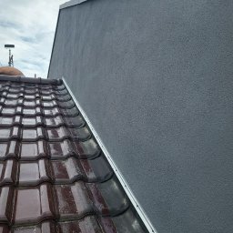 Bartosz Szewczykowski - Szara, świeżo otynkowana ściana domu z widocznym fragmentem mokrego, brązowego dachu z dachówki. Widoczne łączenie ściany z dachem.