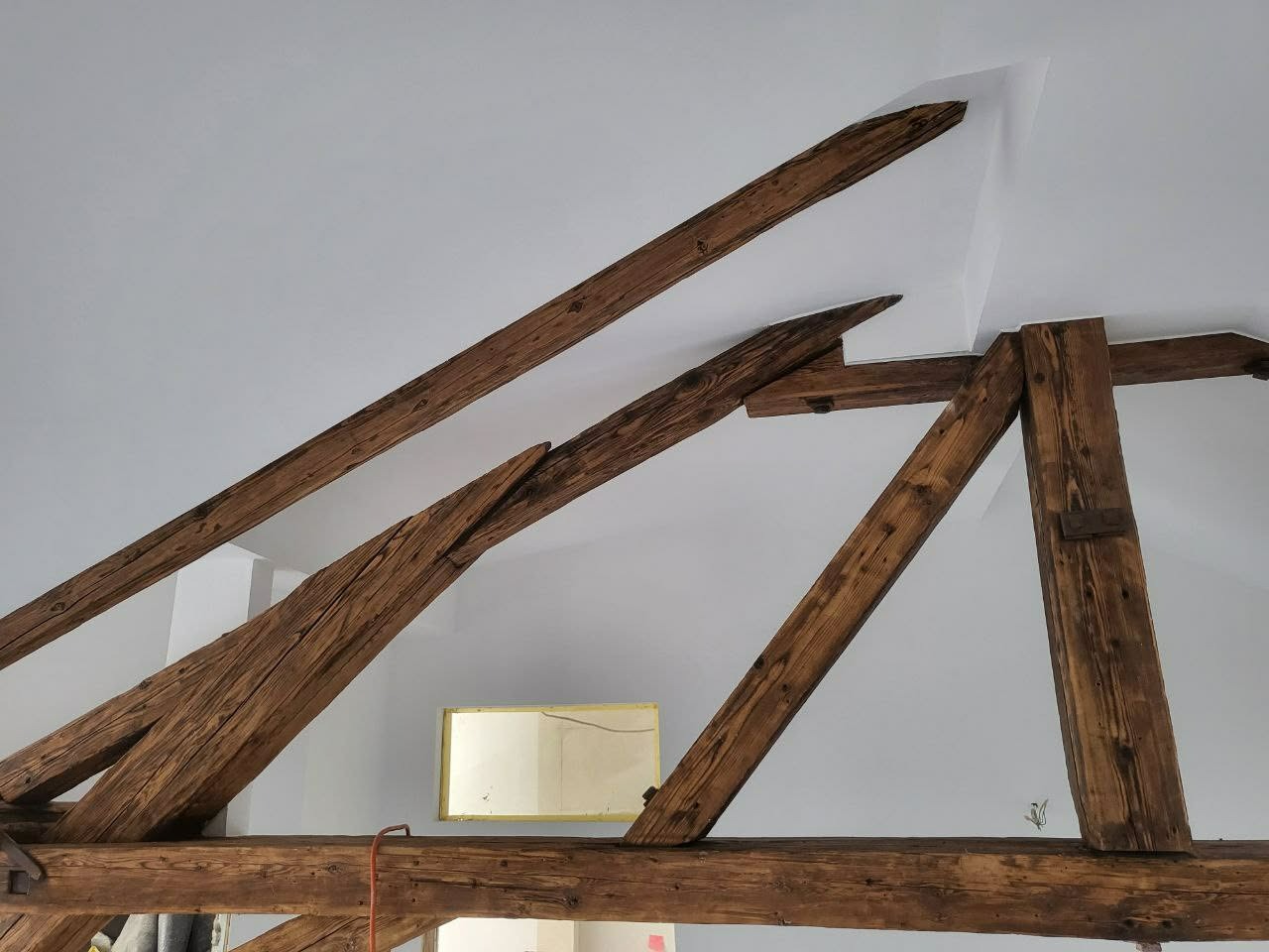 Drewniana konstrukcja więźby dachowej z widocznymi belkami na tle białego sufitu i ściany. Elementy złączone metalowymi okuciami. Widoczny fragment okna.