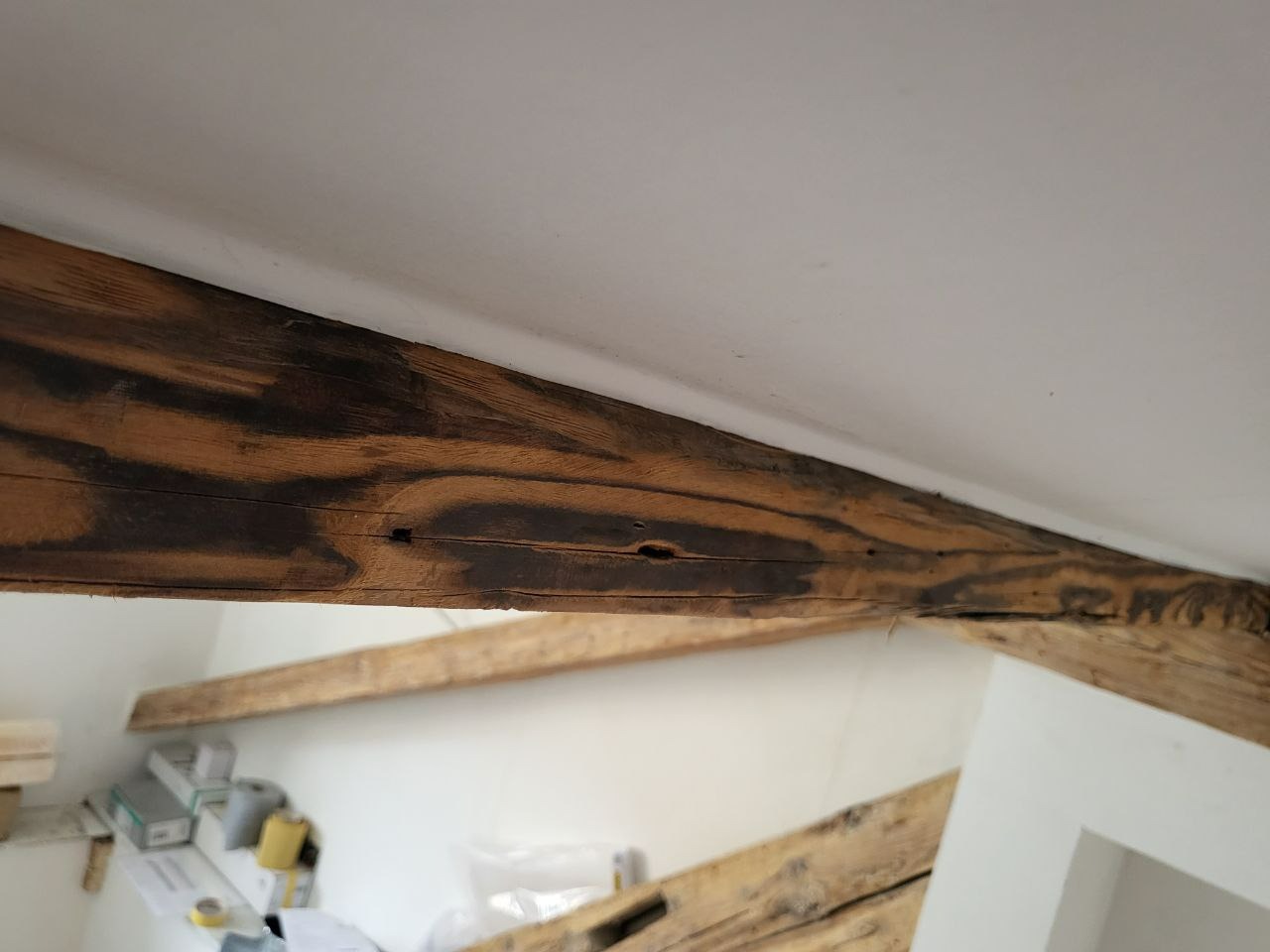 Drewniana belka stropowa z wyraźnym usłojeniem, częściowo pomalowana na ciemny brąz, widoczna na tle białego sufitu i ściany. Element konstrukcyjny w trakcie prac wykończeniowych.