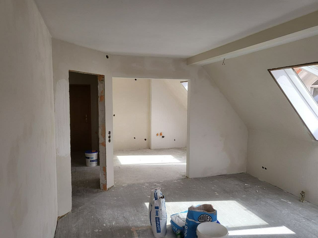 Poddasze po szpachlowaniu, widoczne worki z zaprawą i wiaderko. Ściany przygotowane do dalszych prac wykończeniowych, okno dachowe wpuszcza światło.