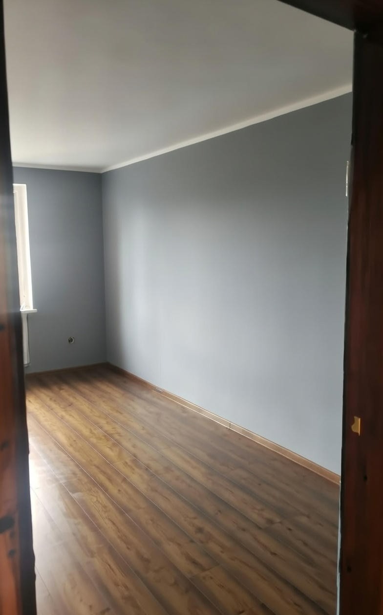 Pomalowane na szaro wnętrze pokoju z panelami podłogowymi, widoczne przez otwarte, drewniane drzwi. Ściany gładkie, sufit biały z listwą. Okno po lewej stronie.