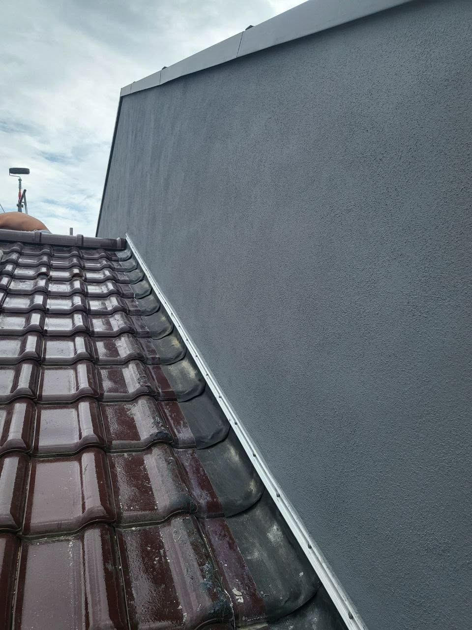 Szara, świeżo otynkowana ściana domu z widocznym fragmentem mokrego, brązowego dachu z dachówki. Widoczne łączenie ściany z dachem.