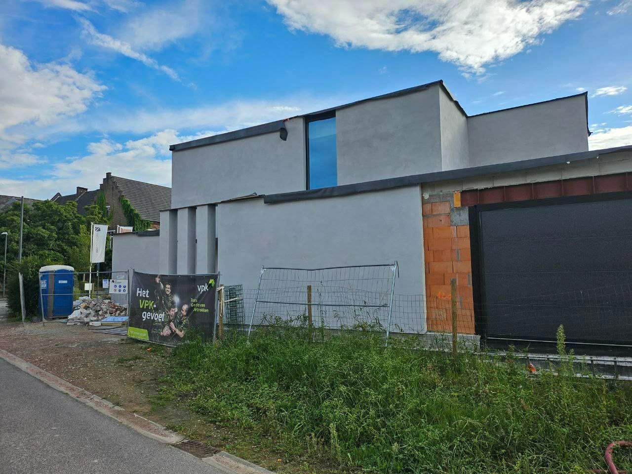 Surowy, szary dom jednorodzinny w trakcie budowy, z widocznymi fragmentami muru z cegły i świeżo otynkowanymi ścianami. Na pierwszym planie trawa i ogrodzenie budowlane.