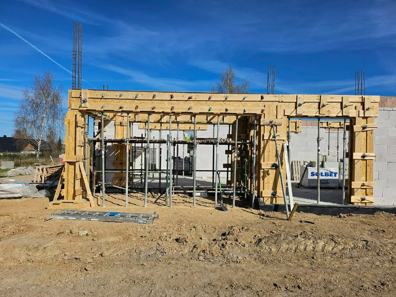 Wznoszenie ścian domu z bloczków Solbet, widoczne drewniane szalunki i rusztowania. Niebo błękitne, w tle drzewa i zabudowania.