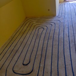 Hydro-Instal Adrian - Niebieskie rury ogrzewania podłogowego ułożone w pętle na szarej wylewce, widok z góry, żółta ściana w tle. Instalacja przed zalaniem posadzki.