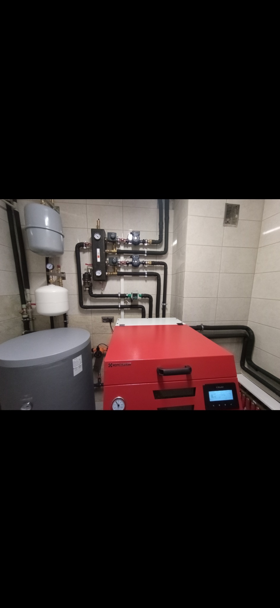 Nowoczesna kotłownia z czerwonym piecem Kotłospaw, zbiornikiem buforowym i rozbudowaną instalacją hydrauliczną na tle jasnych, kwadratowych płytek.