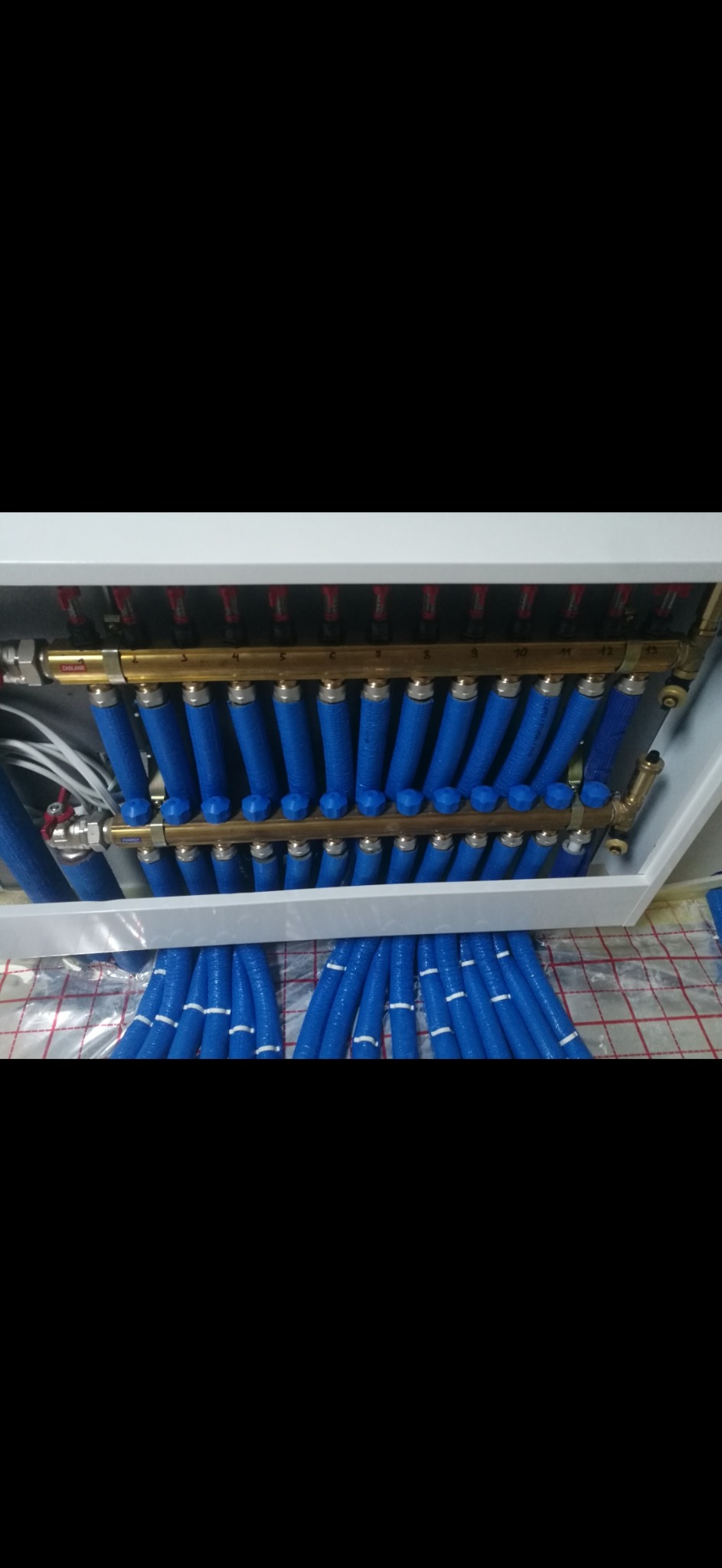 Rozdzielacz ogrzewania podłogowego z niebieskimi rurami w izolacji, widoczne zawory i numeracja. Instalacja na folii izolacyjnej z nadrukiem siatki.