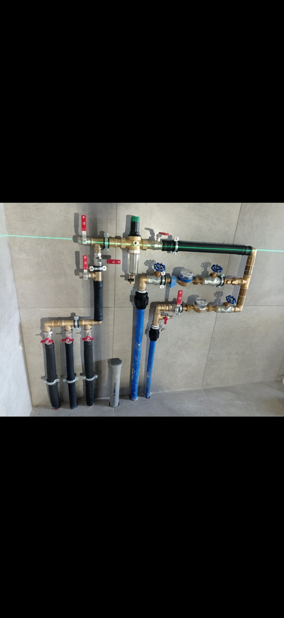 Złożona instalacja wodna z rur miedzianych, zaworów i filtrów, zamontowana na szarej ścianie z płytek. Widoczna zielona linia lasera poziomującego.