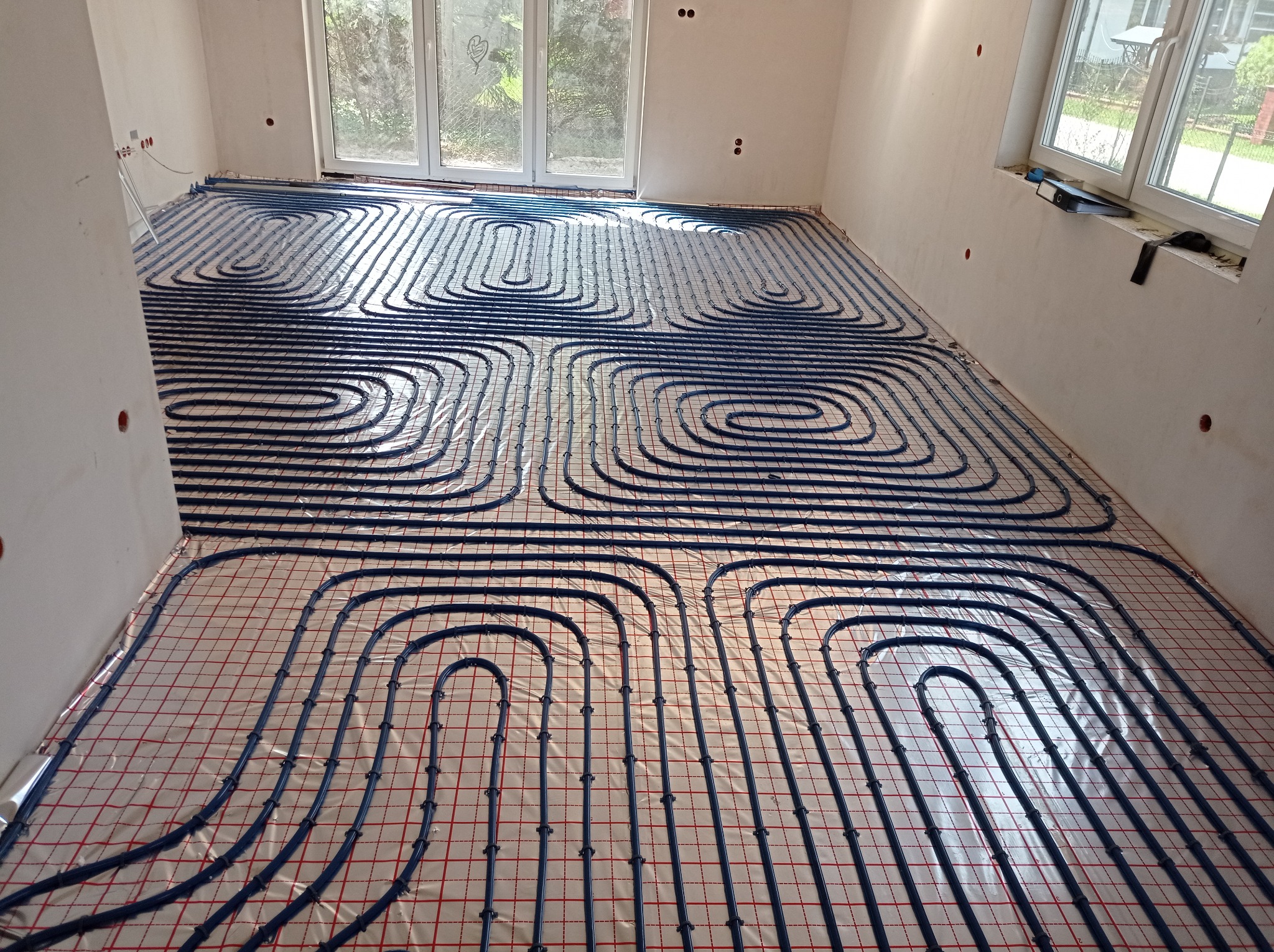 Ułożone, niebieskie rury ogrzewania podłogowego na folii z czerwoną siatką w jasnym pomieszczeniu z oknem. Widok z góry, etap instalacji przed wylewką.