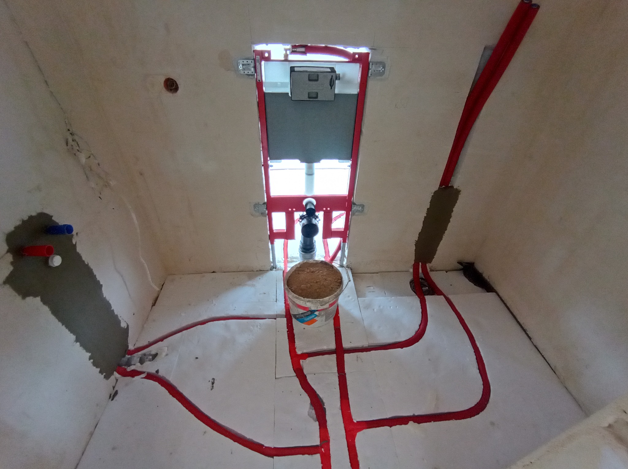 Instalacja ogrzewania podłogowego z czerwonymi rurami na białym styropianie, widok z góry. Częściowo zamontowany stelaż podtynkowy WC.