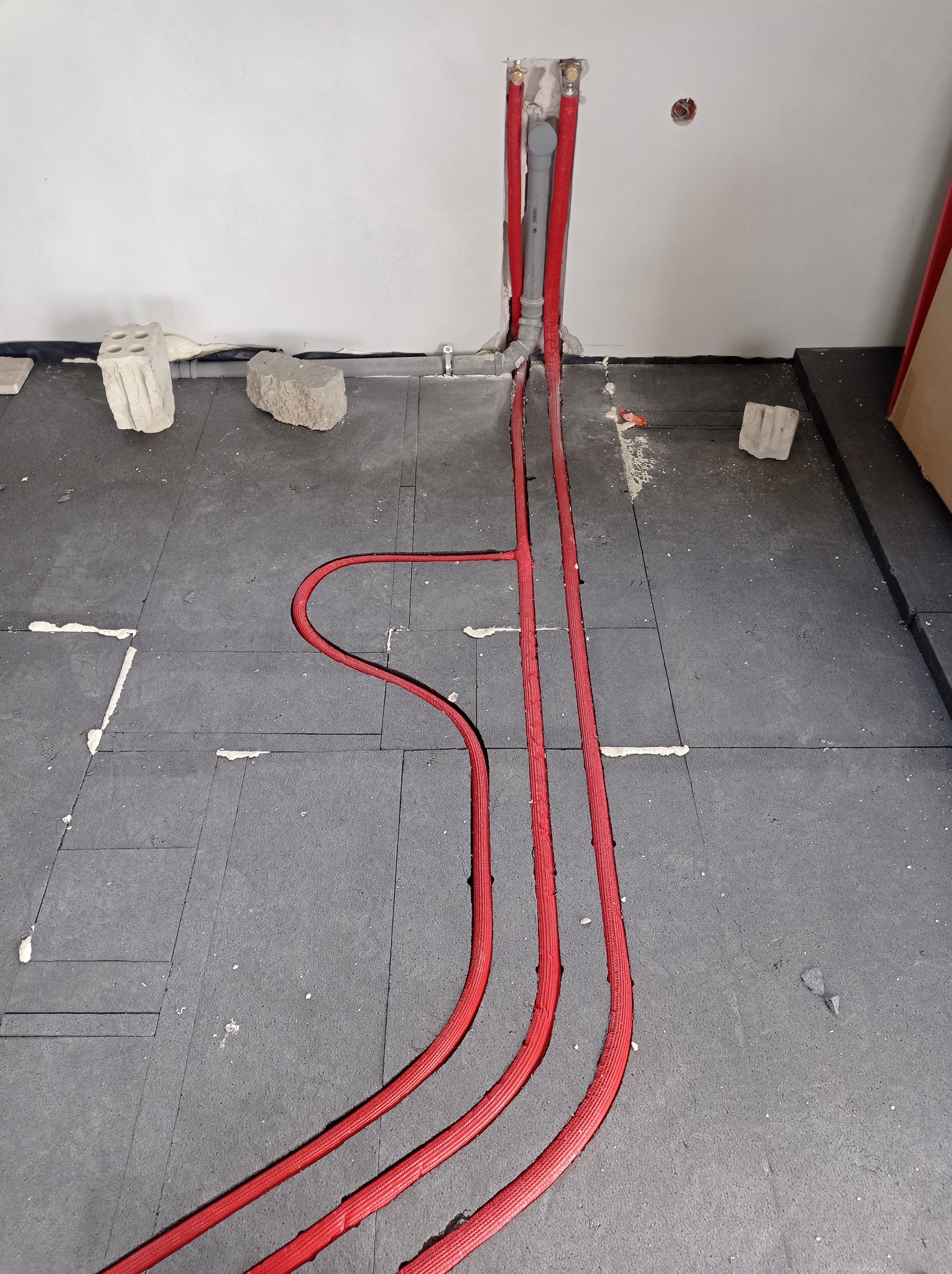 Czerwone rury w izolacji ułożone w bruzdach na czarnej macie podłogowej, prawdopodobnie instalacja ogrzewania podłogowego w trakcie montażu.