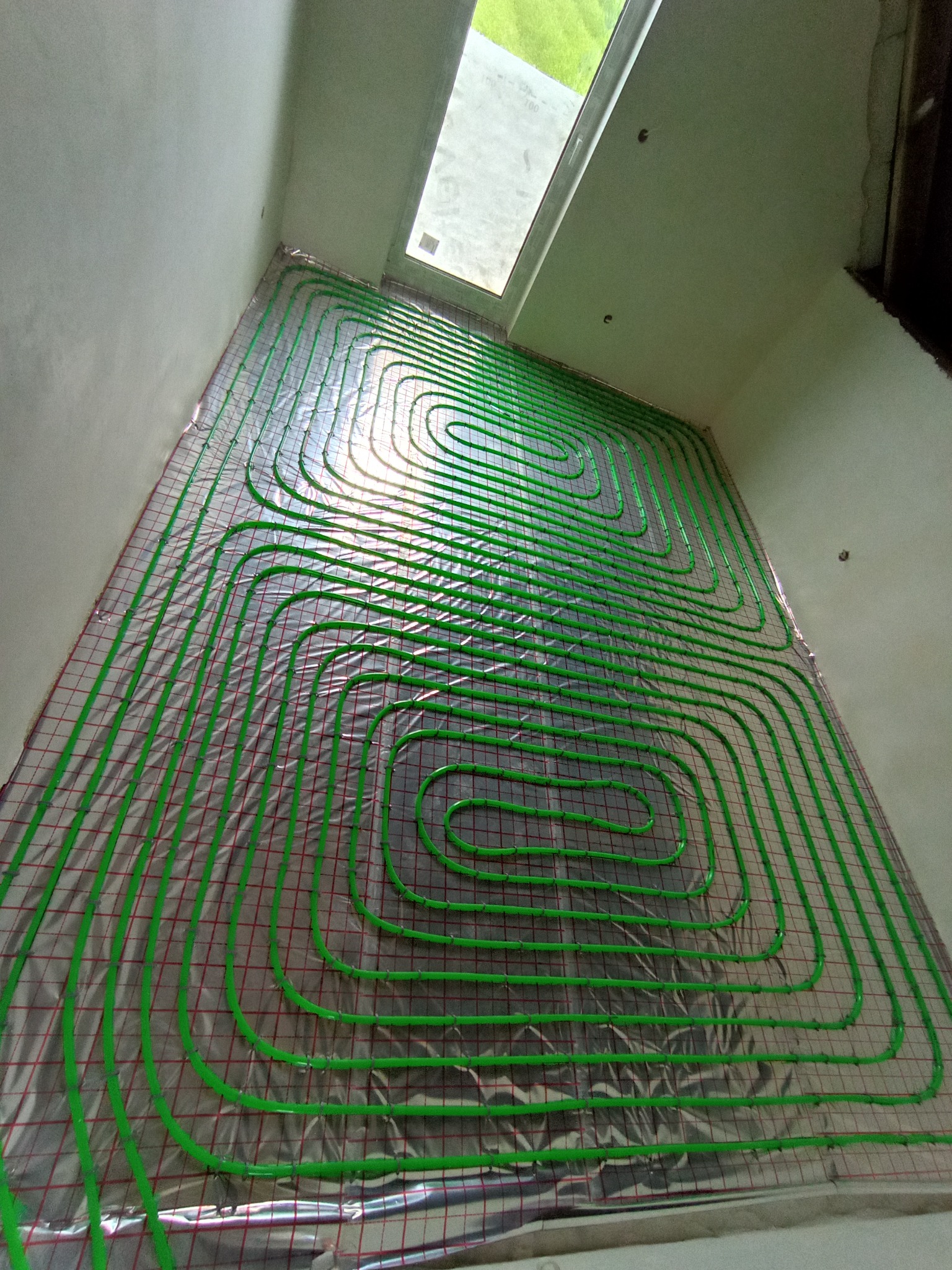 Zielone rury ogrzewania podłogowego ułożone na srebrnej folii z czerwoną siatką, widok z góry, w tle okno dachowe w surowym wnętrzu.