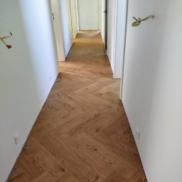 KING-FLOOR MIKOŁAJ CHMIEL - Korytarz z parkietem ułożonym w jodełkę francuską. Jasne ściany, widoczne gniazdka elektryczne. W tle otwarte drzwi do kolejnych pomieszczeń.