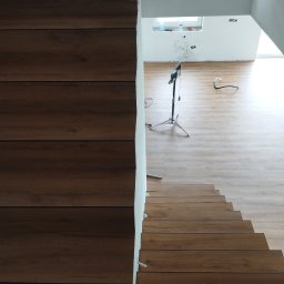 KING-FLOOR MIKOŁAJ CHMIEL - Ukończone schody z paneli drewnopodobnych, widok z góry. Na podłodze widoczny statyw oświetleniowy i przewody. Świeżo wykończone wnętrze.