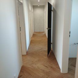 KING-FLOOR MIKOŁAJ CHMIEL - Korytarz z parkietem ułożonym w jodełkę. Widoczne białe ściany, drzwi z czarnymi skrzydłami i punktowe oświetlenie sufitowe. Perspektywa zbieżna w głąb.