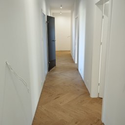KING-FLOOR MIKOŁAJ CHMIEL - Korytarz z parkietem ułożonym w jodełkę, białe ściany i drzwi, widoczne niedokończone prace elektryczne. Jasne wnętrze z nowoczesnym oświetleniem.