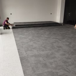 KING-FLOOR MIKOŁAJ CHMIEL - Montaż szarej wykładziny dywanowej w dużym pomieszczeniu z podestem. Pracownik docina wykładzinę przy ścianie. Część podłogi wyłożona jasną wykładziną.