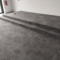 KING-FLOOR MIKOŁAJ CHMIEL - Nowoczesna, dwustopniowa podłoga wyłożona szarą wykładziną dywanową w nowym budynku. Widoczny fragment ściany i okna. Wykończenie listwami.