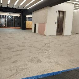 KING-FLOOR MIKOŁAJ CHMIEL - Jasna wykładzina dywanowa w biurze, wzór geometryczny, widoczne zabezpieczenia krawędzi taśmą. W tle ściany i winda w surowym stanie.