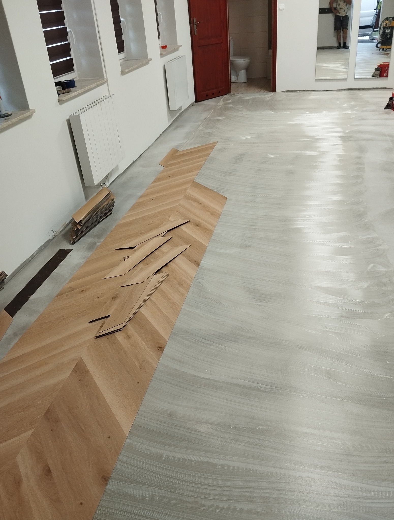 Układanie parkietu w jodełkę: widok z góry na częściowo ułożoną podłogę, z przygotowanymi panelami i podkładem. W tle okna i drzwi do łazienki.